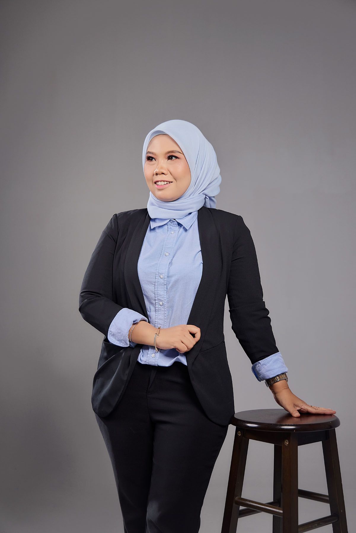 Stylish Muslim woman in blue hijab and black blazer posing beside a wooden stool in studio.