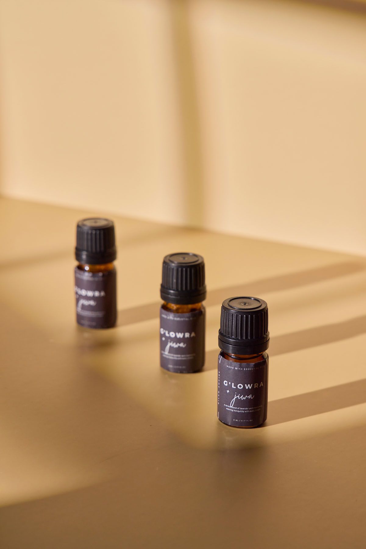 Three amber glass C'LOWRA Jiwa essential oil bottles arranged on a golden beige surface with soft shadows.