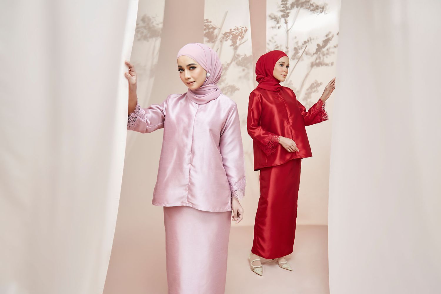 Two women modeling pink and red satin baju kurung sets with matching hijabs in a bright studio backdrop.