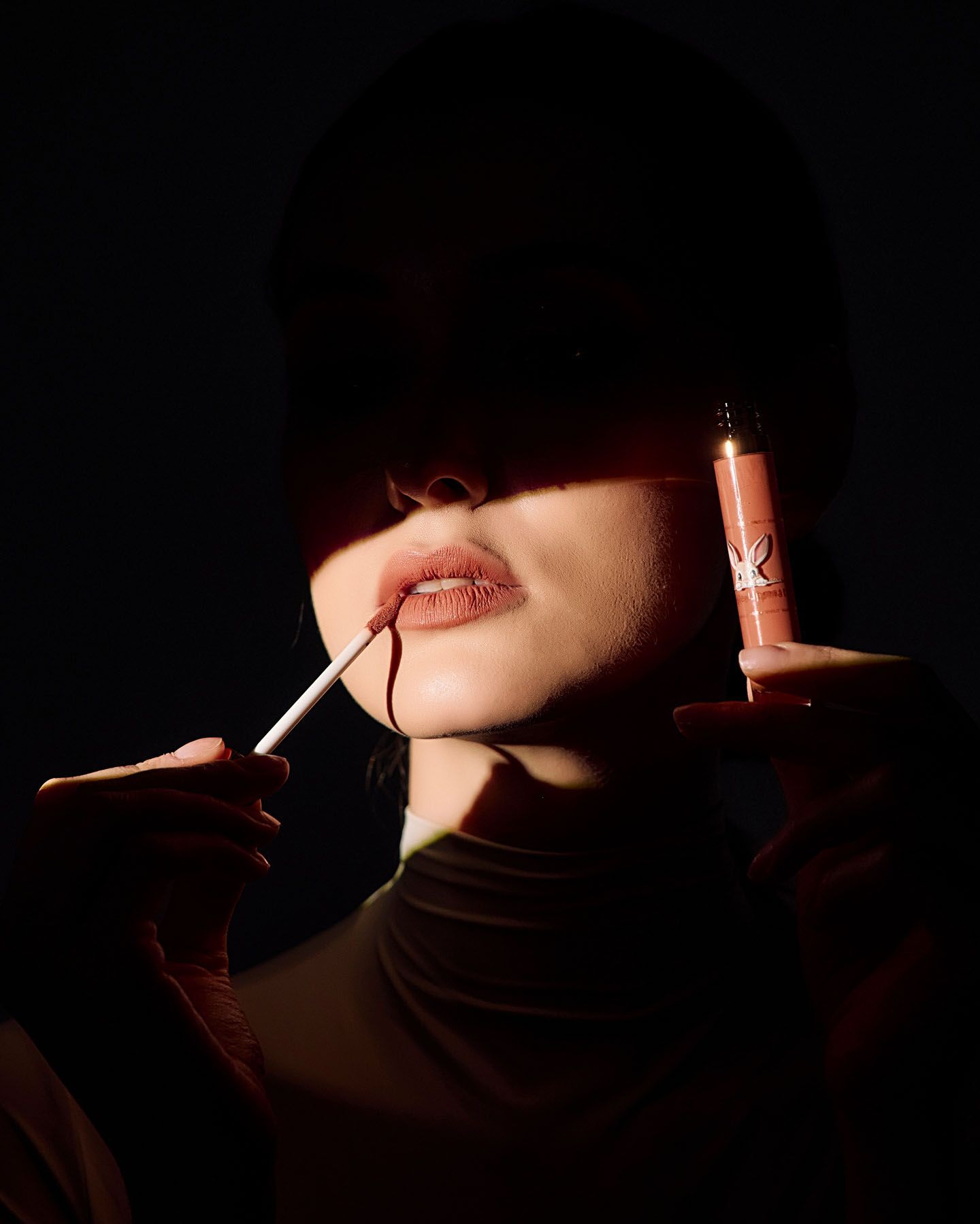 Close-up of woman applying nude lip gloss in dramatic dark studio lighting.