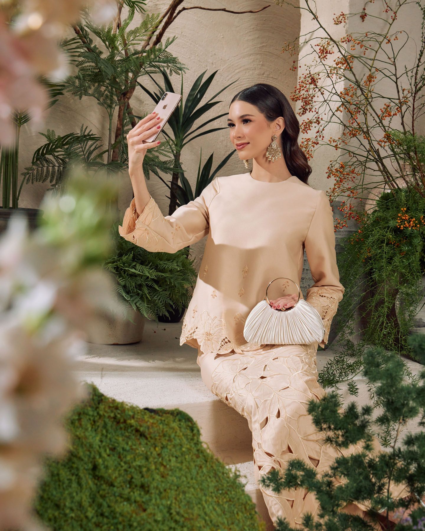 Elegant woman in beige two-tone baju kurung taking selfie while holding white clutch bag outdoors.