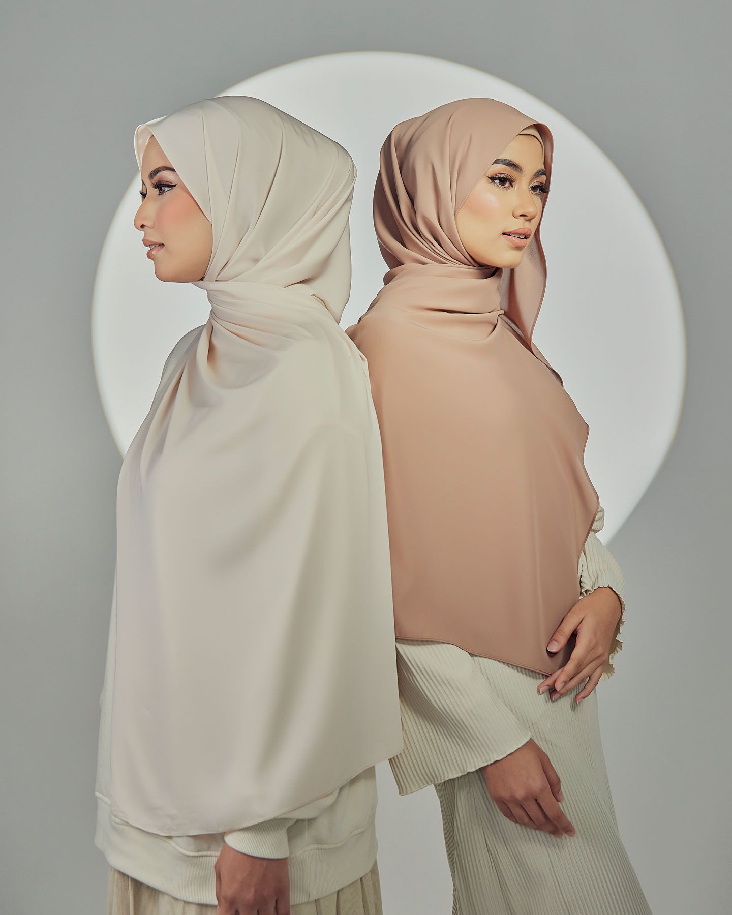 Two women in elegant cream and beige hijabs posed back-to-back against a minimalist grey background.