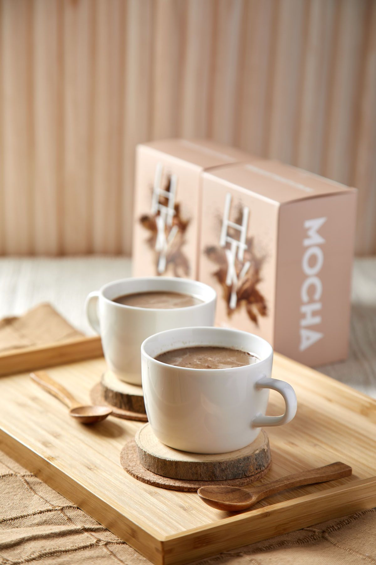 Two white cups of mocha coffee on wooden coasters with spoons on a bamboo tray beside mocha brand boxes.