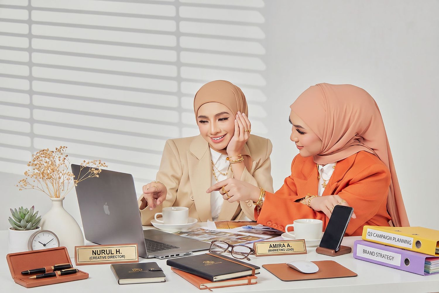 Two Muslim women in hijabs collaborating at a desk with laptop in a bright office setting.