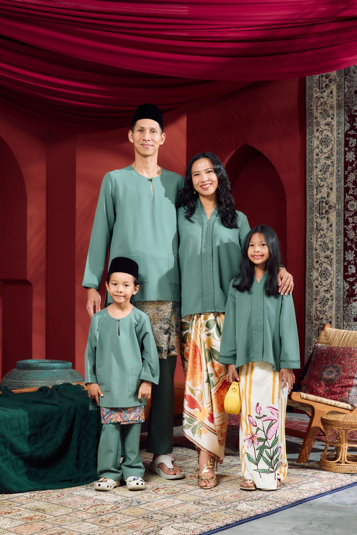 Family of four in matching sage green Baju Melayu and Baju Kurung standing in a red Moroccan-style set.