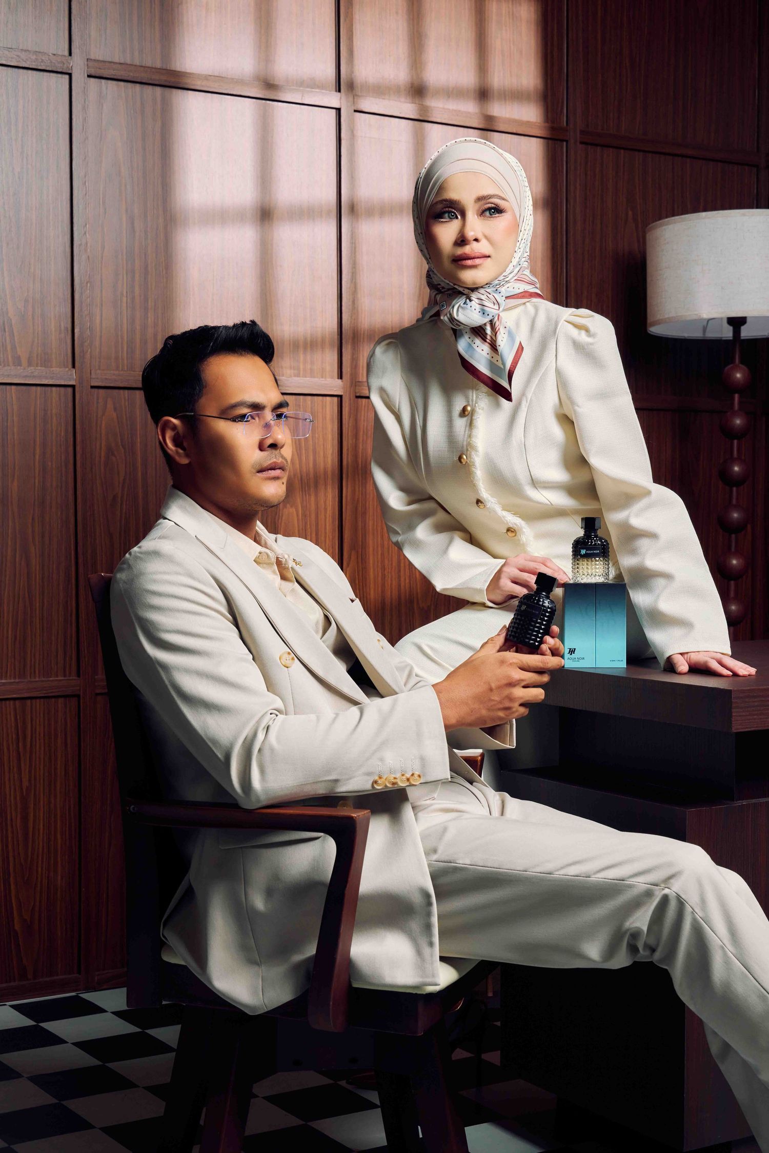 Stylish couple in matching cream outfits posing with fragrance bottle in a vintage wood-paneled room.