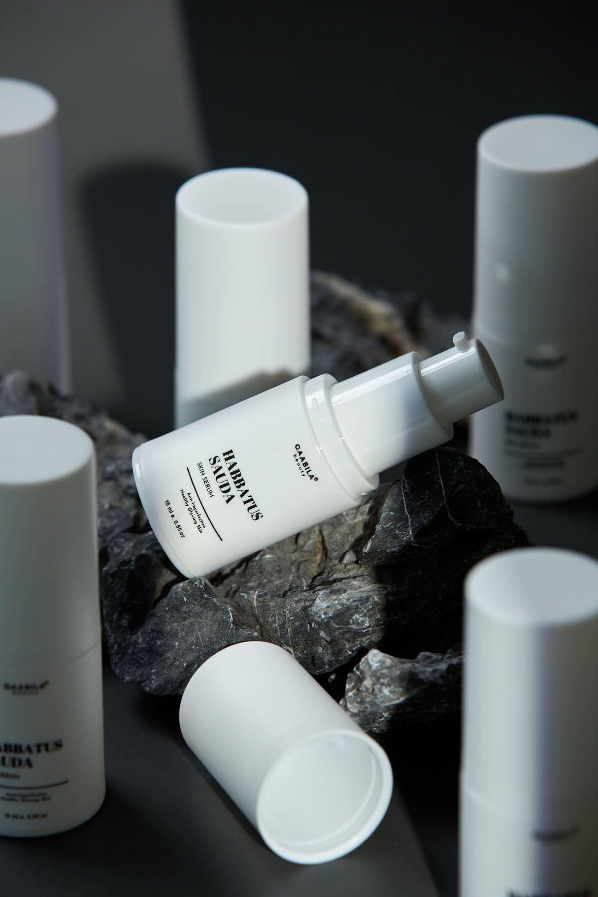 White Habbatus Sauda skincare pump bottles arranged on dark rocks against a moody dark gray background.