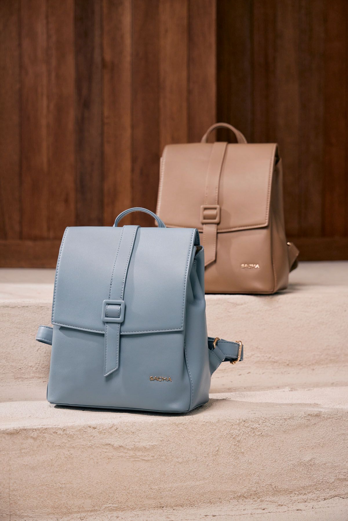 Two structured leather backpacks in blue and tan displayed on stone steps against a wooden backdrop.