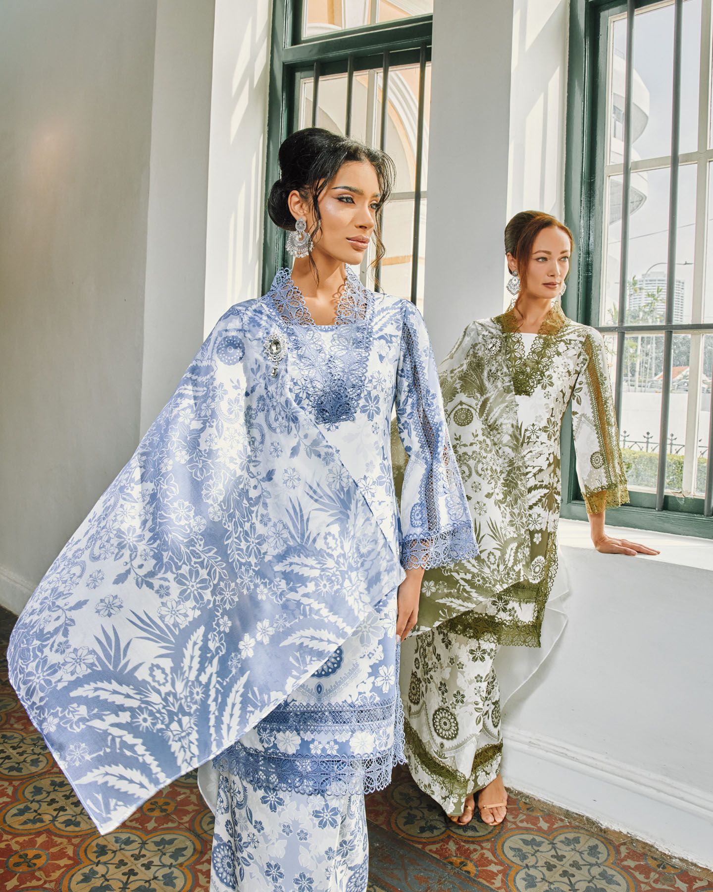 Two women in blue and olive floral baju kurung with cape sleeves in a colonial hallway.