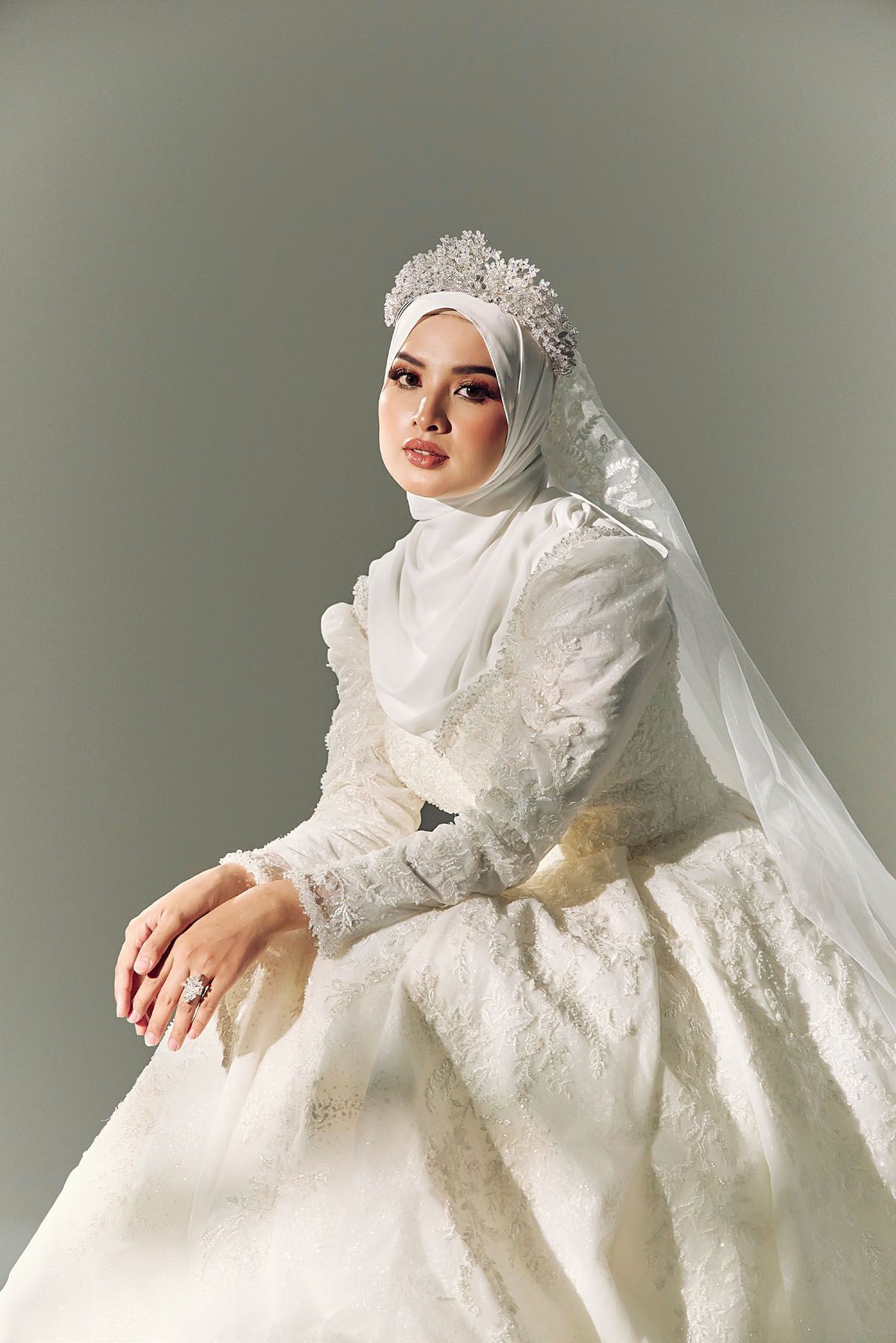 Muslim bride in a white hijab with floral crown and lace wedding dress seated gracefully.