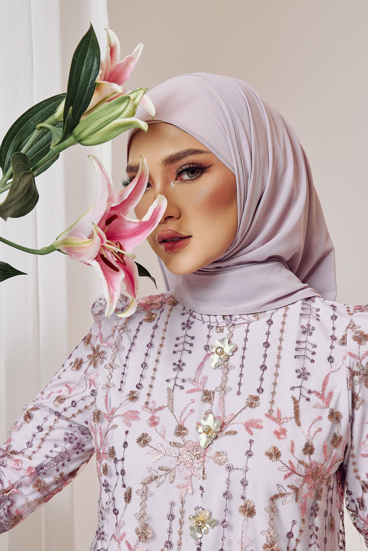 Close-up portrait of a Muslim woman in lavender hijab and floral embroidered dress holding pink lily flowers.