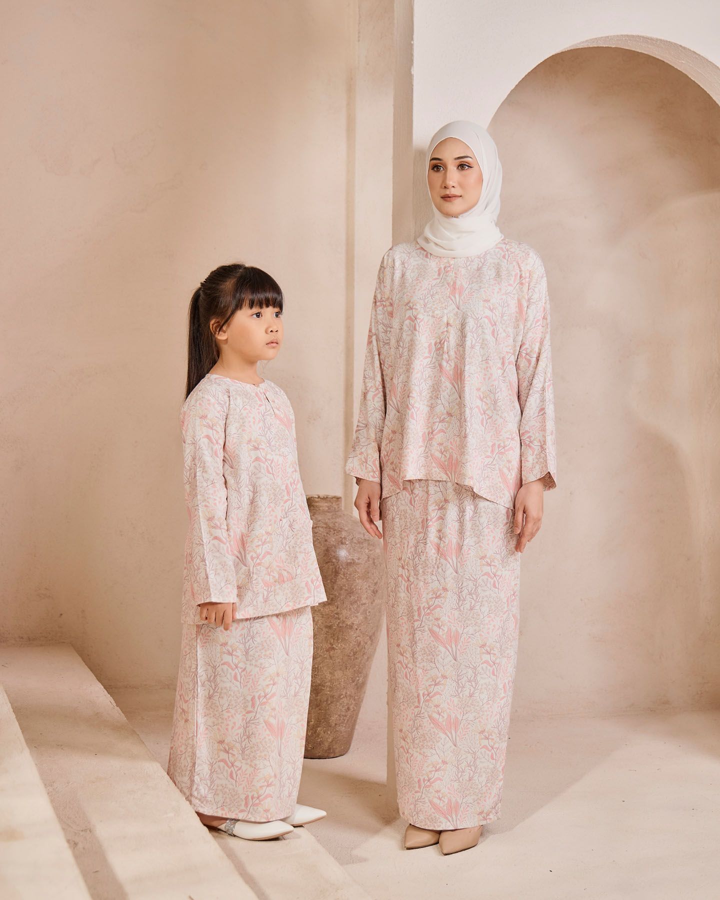 A mother and daughter wear matching pink floral baju kurung outfits, posing together in a soft beige studio.