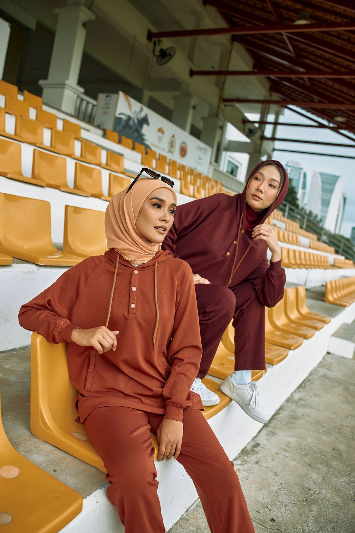 Two women in matching rust-colored hooded tracksuits pose on stadium bleacher seats.