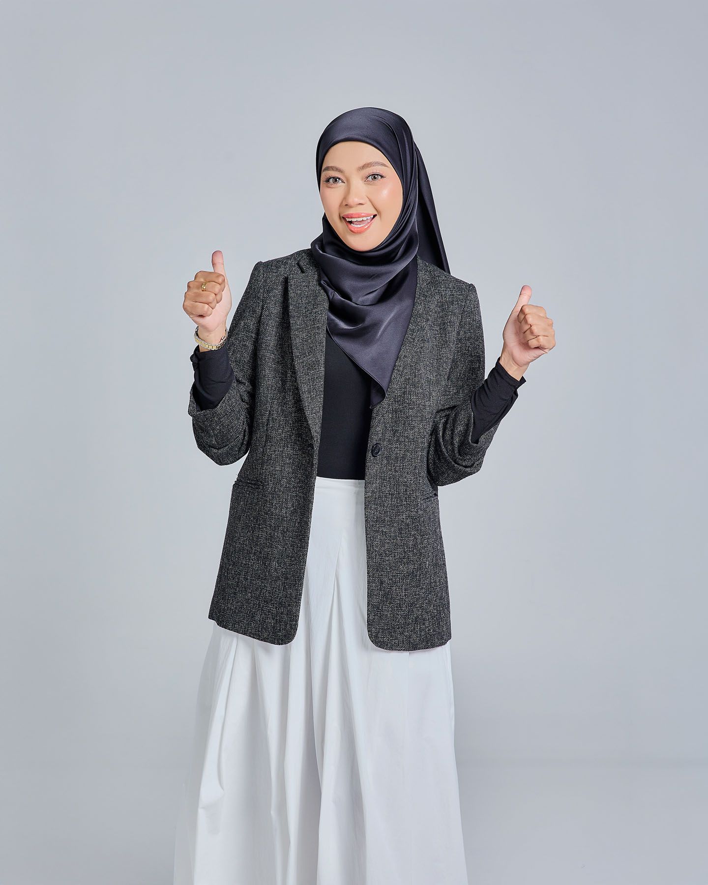 Muslim woman in gray herringbone blazer and navy hijab giving double thumbs up with a big smile.