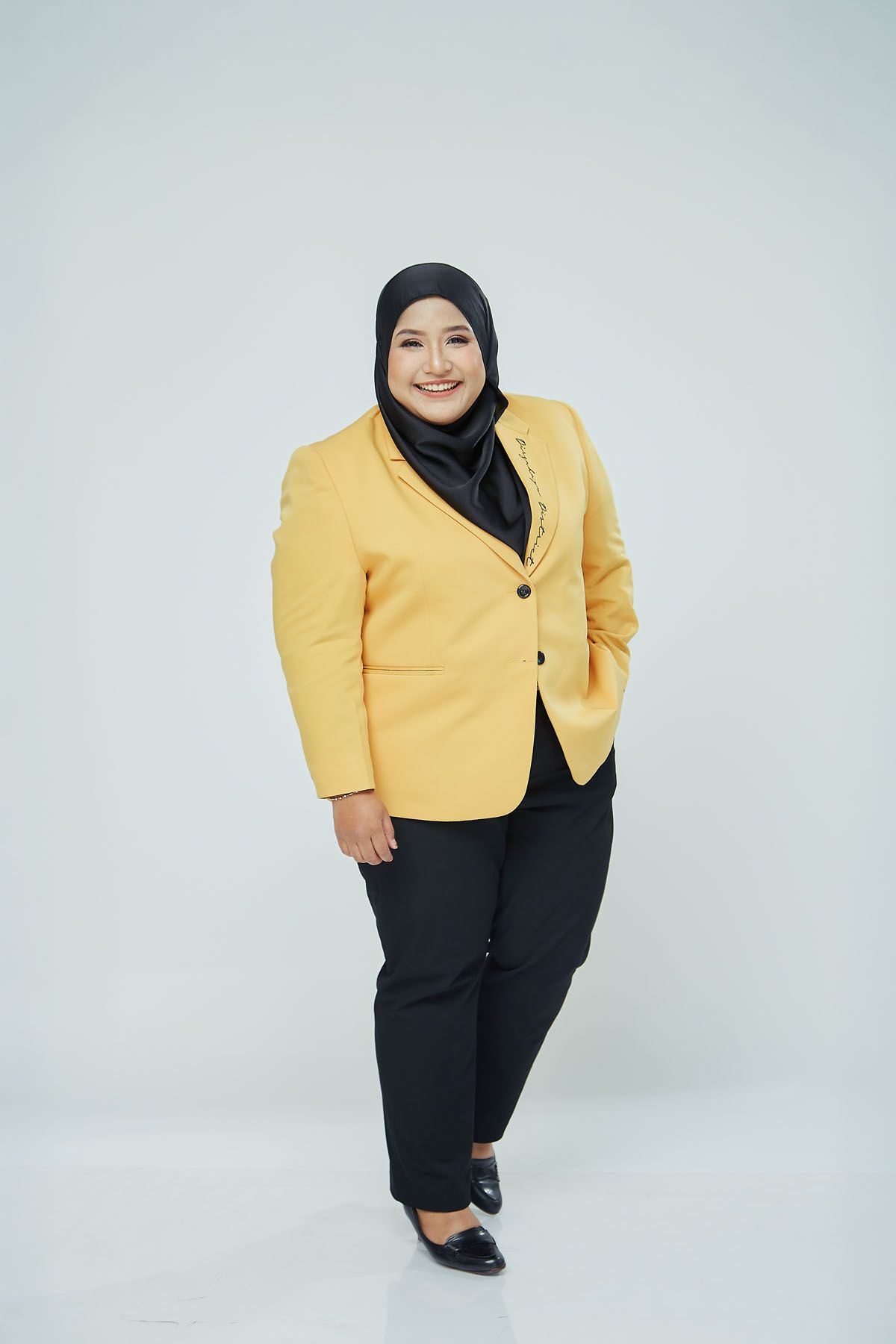Plus-size Muslim woman in yellow blazer and black hijab smiling confidently against white background.