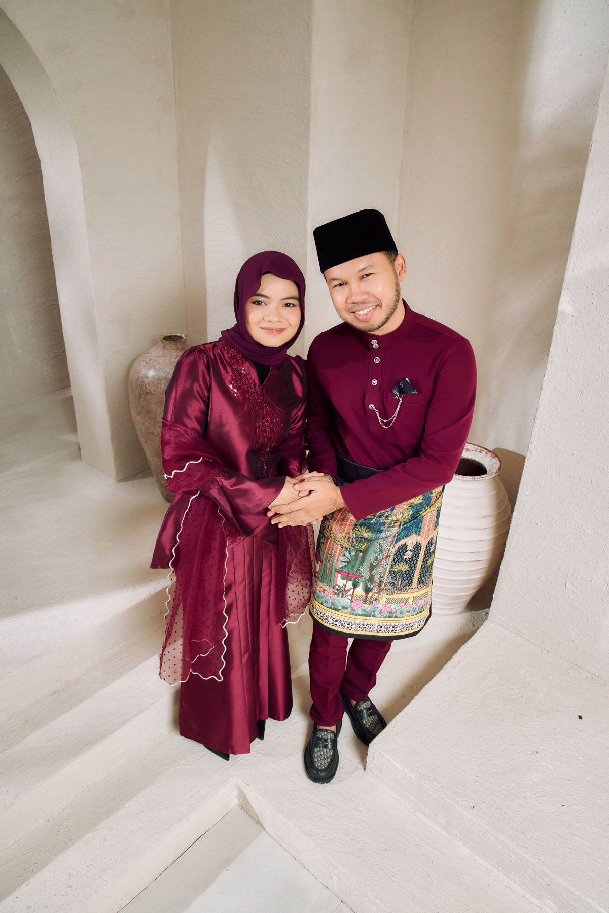 A smiling Malaysian couple in matching maroon traditional Eid outfits sitting closely together on white steps indoors.