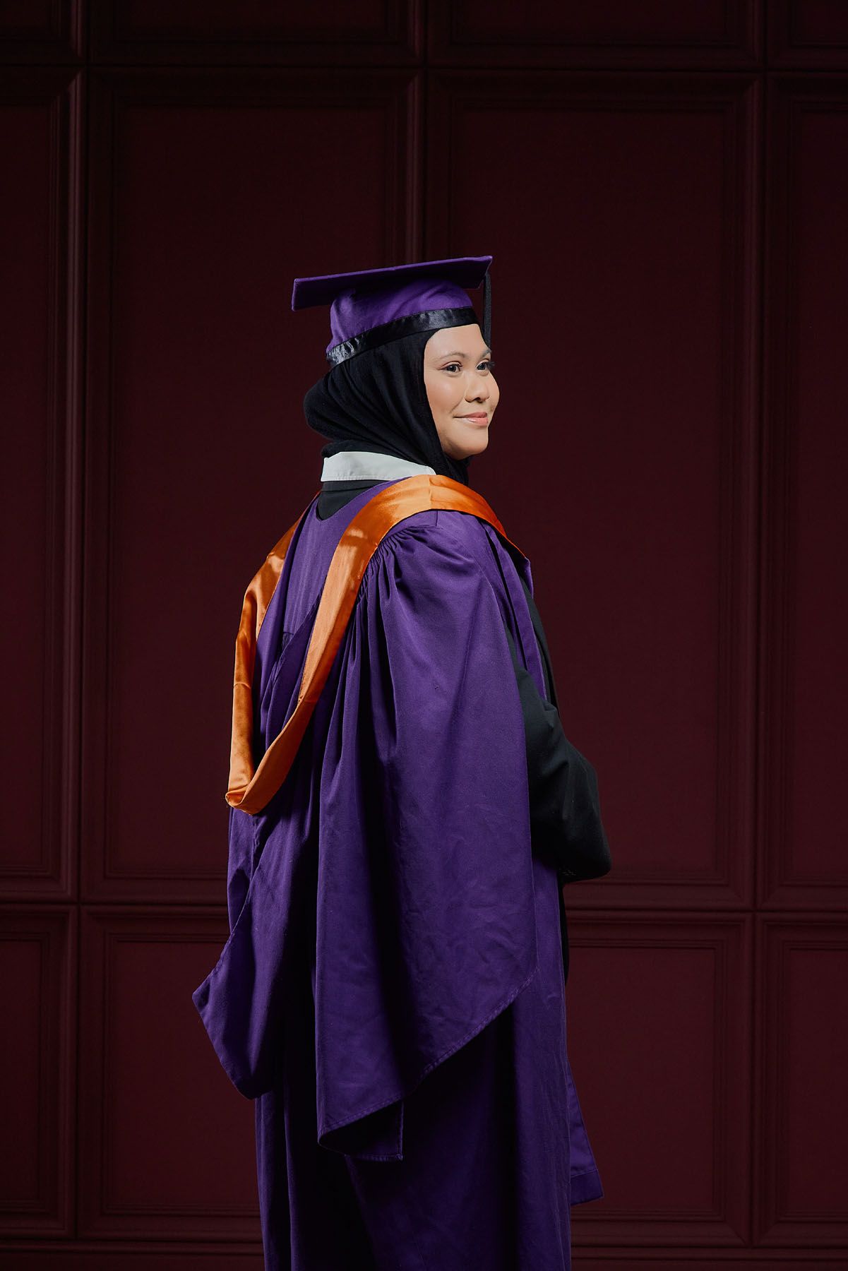 Muslim woman in purple graduation gown and cap with orange sash posing against dark red wall.