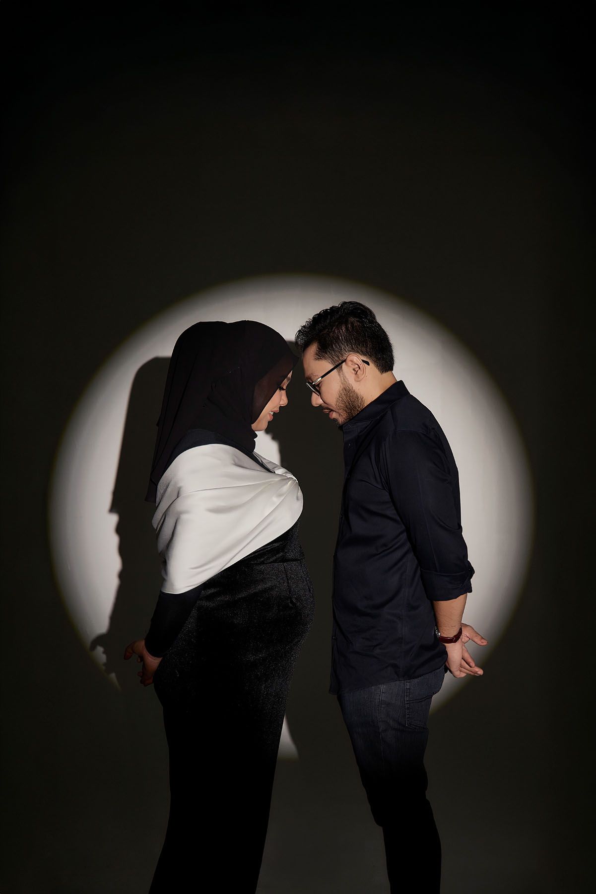 Muslim couple in black attire sharing an intimate moment in a dramatic spotlight on dark background.