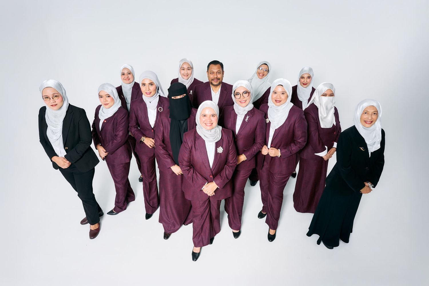 Group of women in matching maroon hijabs and suits posing together with one man in a professional studio setting.