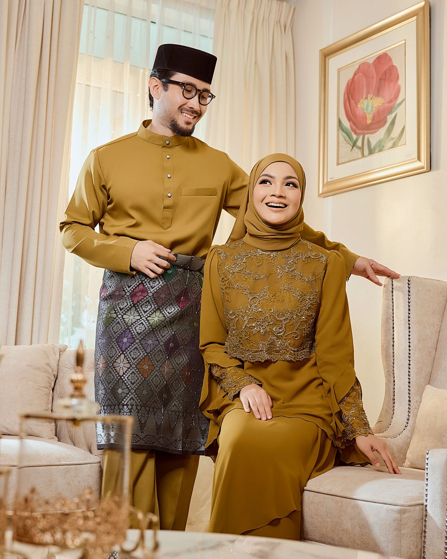 Couple in matching mustard yellow traditional Malay baju kurung and baju melayu outfits posing in elegant living room.