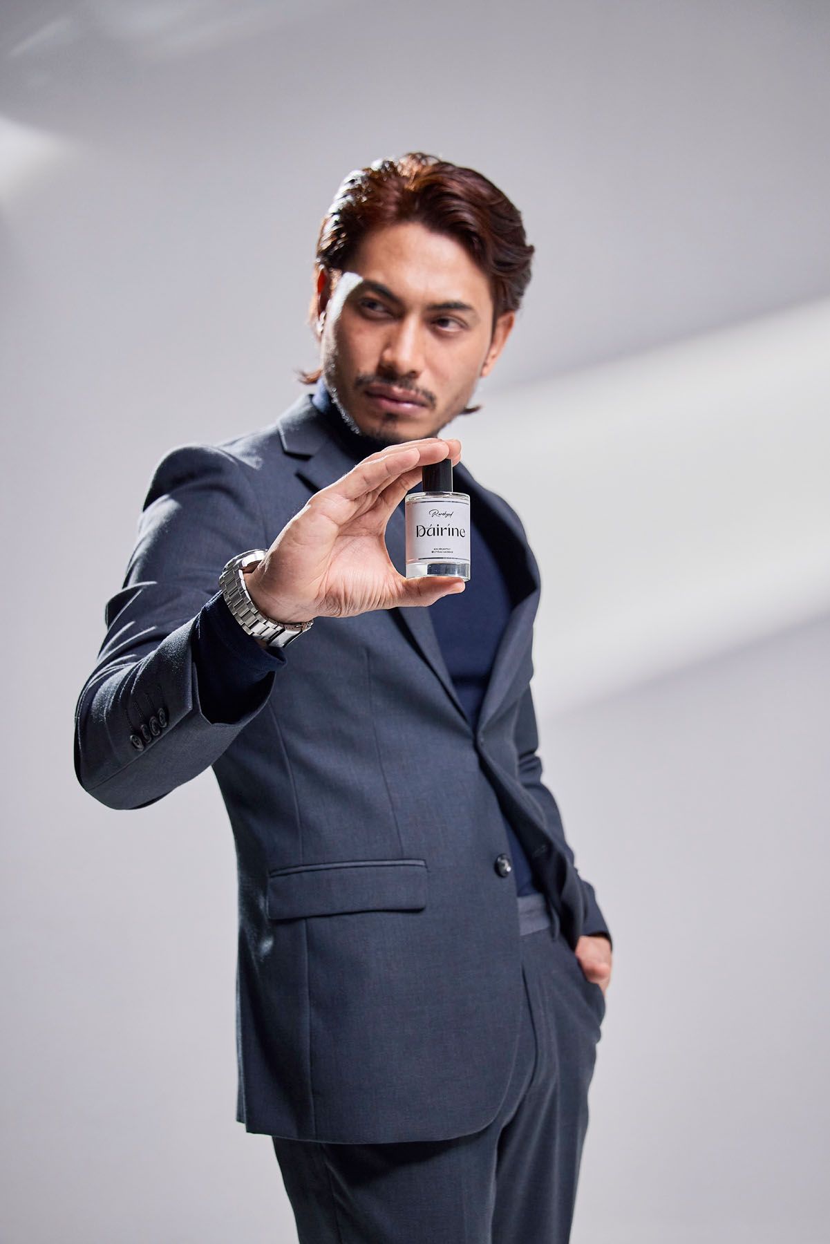 Confident man in grey suit extending hand holding small perfume bottle toward camera in studio.