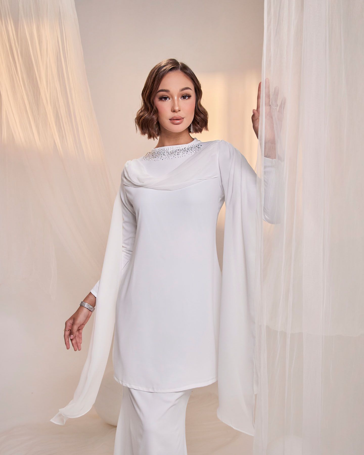 Woman in elegant white long-sleeve tunic with embellished neckline posing with sheer drapes.