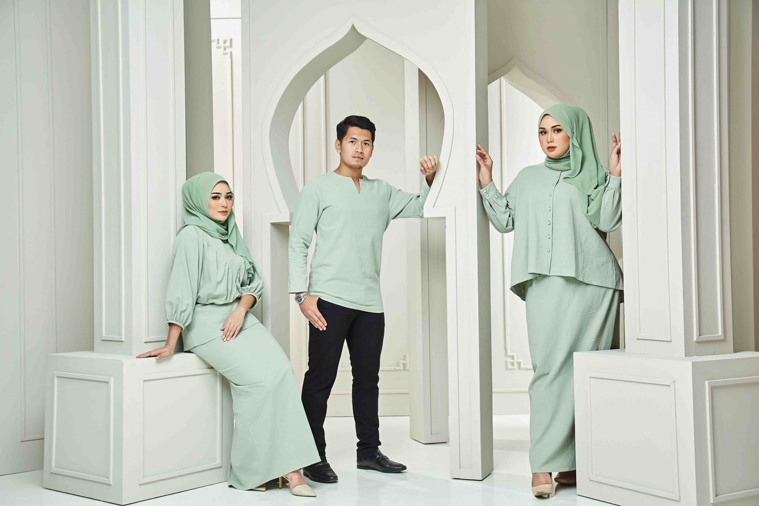 Three models in matching sage green modest outfits with hijabs posing in white architectural studio setting.