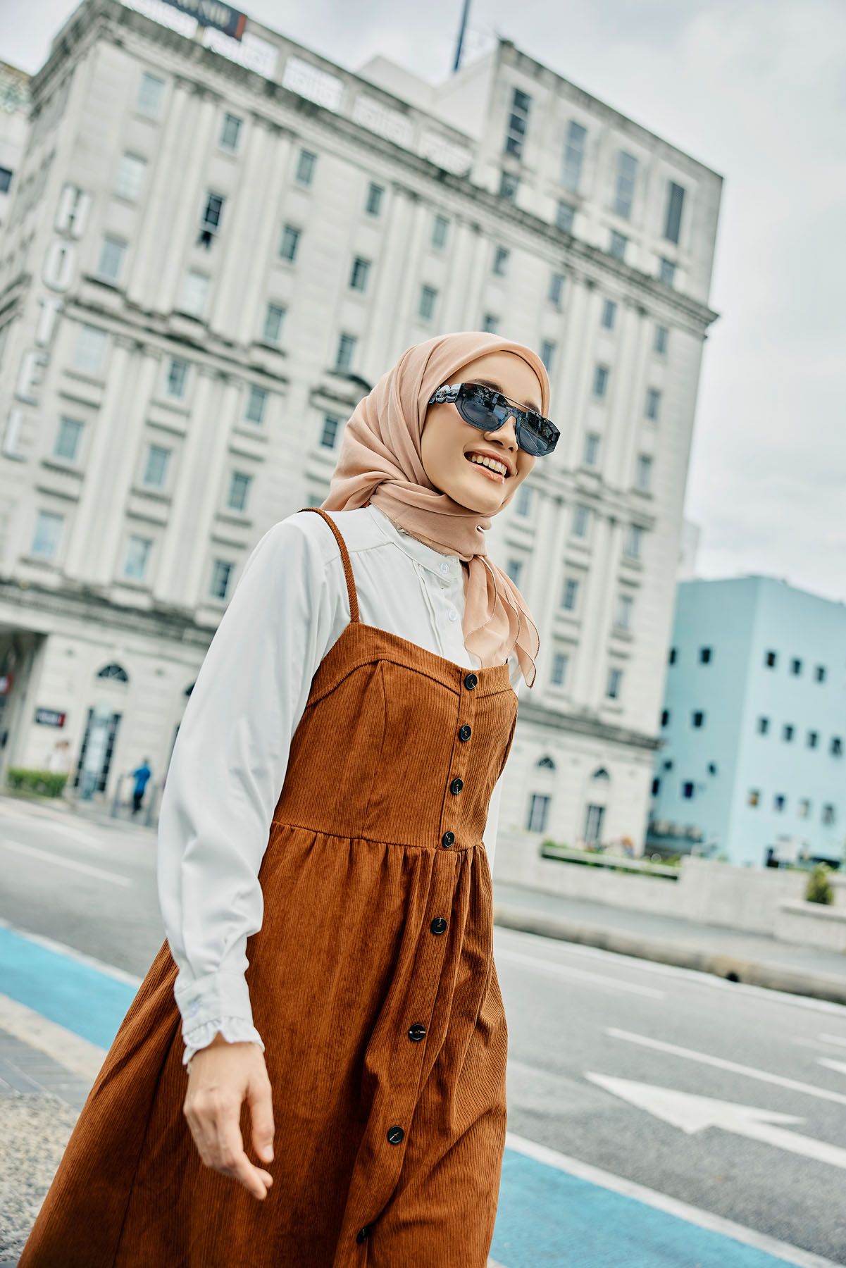 Smiling woman in peach hijab and brown corduroy dress with sunglasses crossing street.