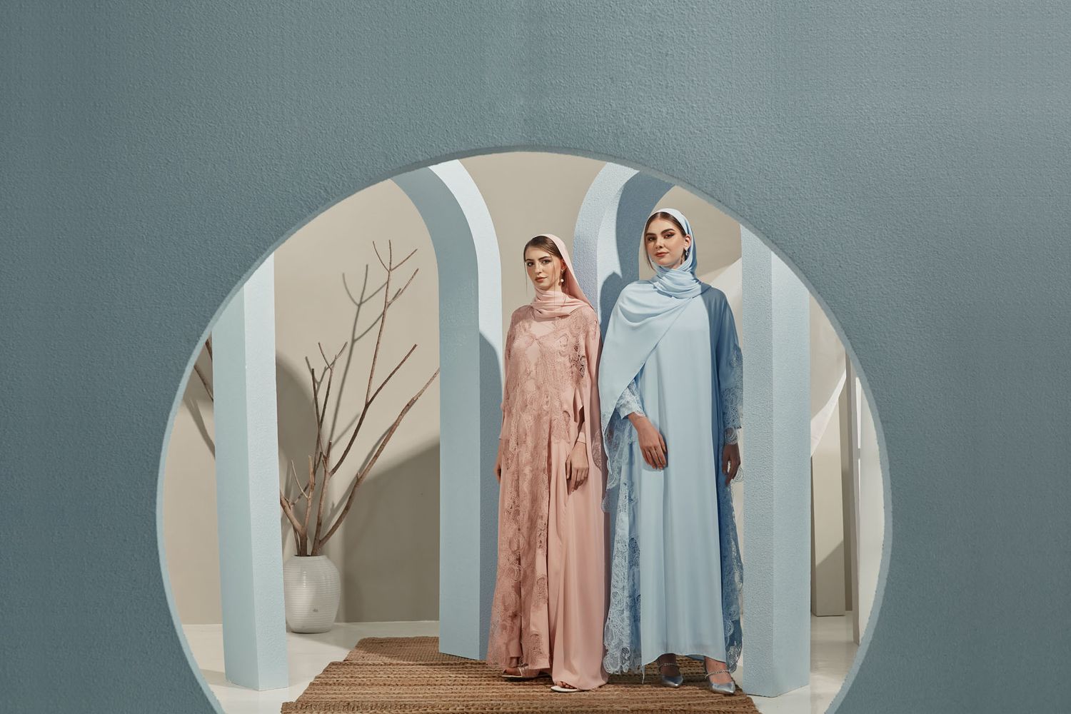 Two women wearing pastel pink and blue lace abayas with hijabs framed in circular arch doorway.