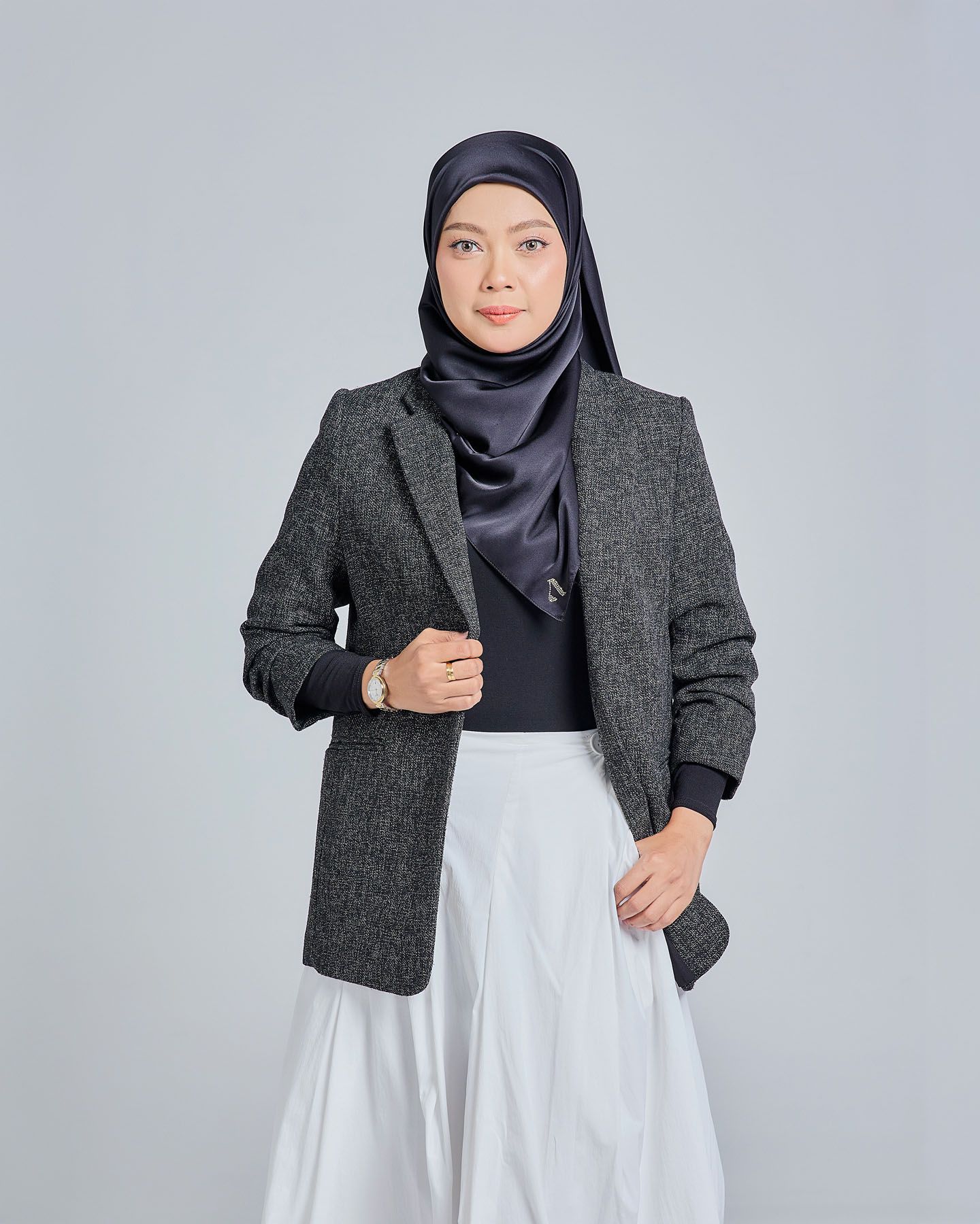 Muslim woman in gray herringbone blazer, black top, white skirt, and navy satin hijab posing confidently.