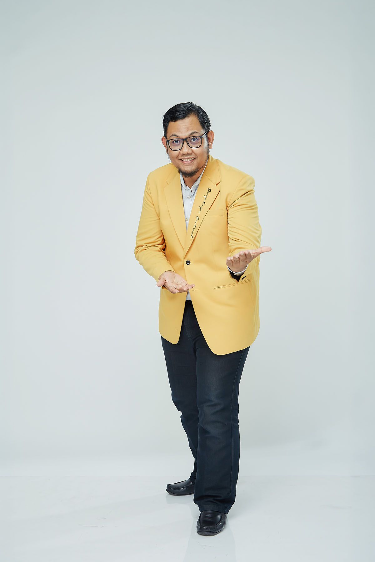 Asian man in a yellow blazer smiling and gesturing with open hands in a white studio setting.