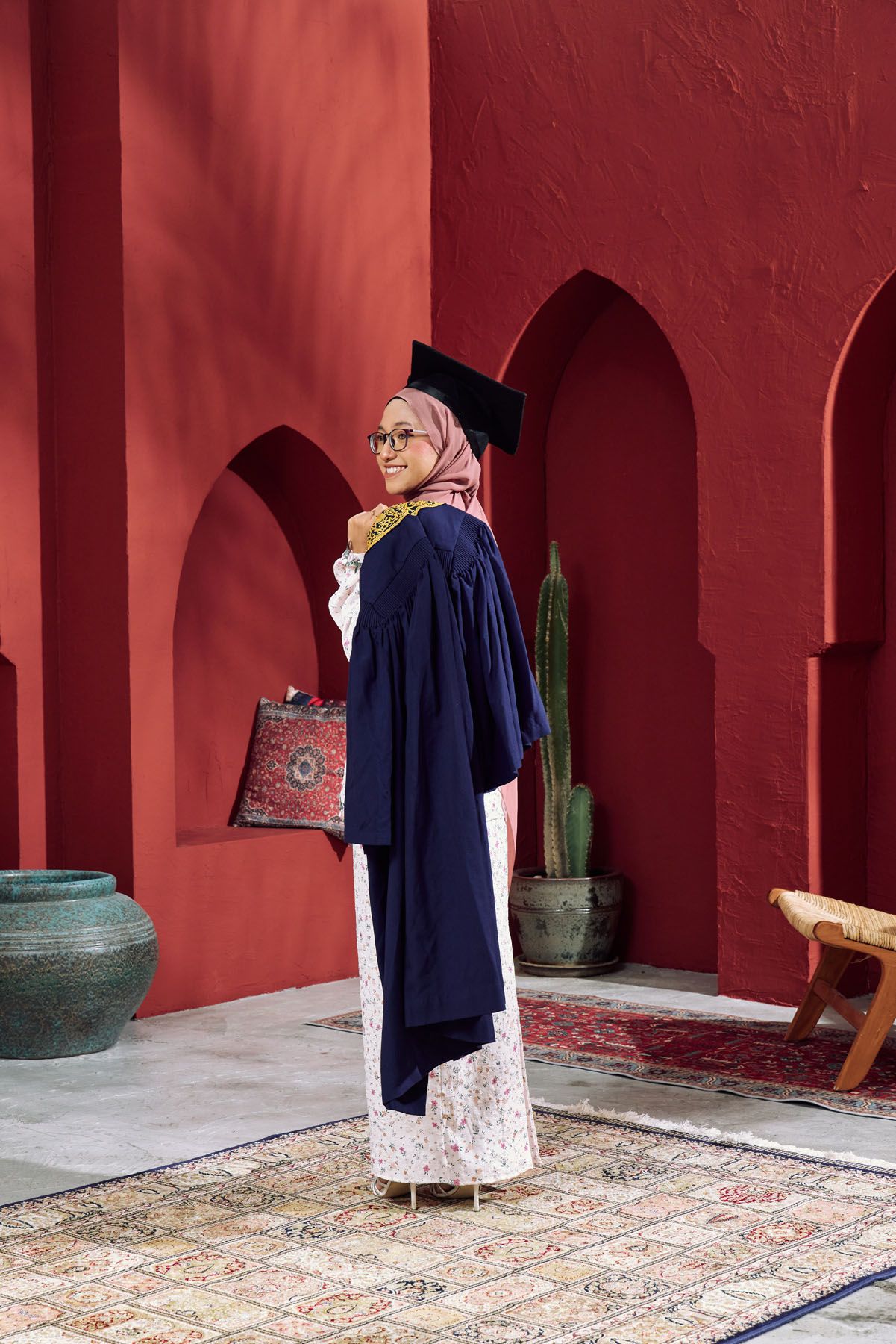 Muslim woman in navy graduation gown and hijab poses in red arched courtyard.