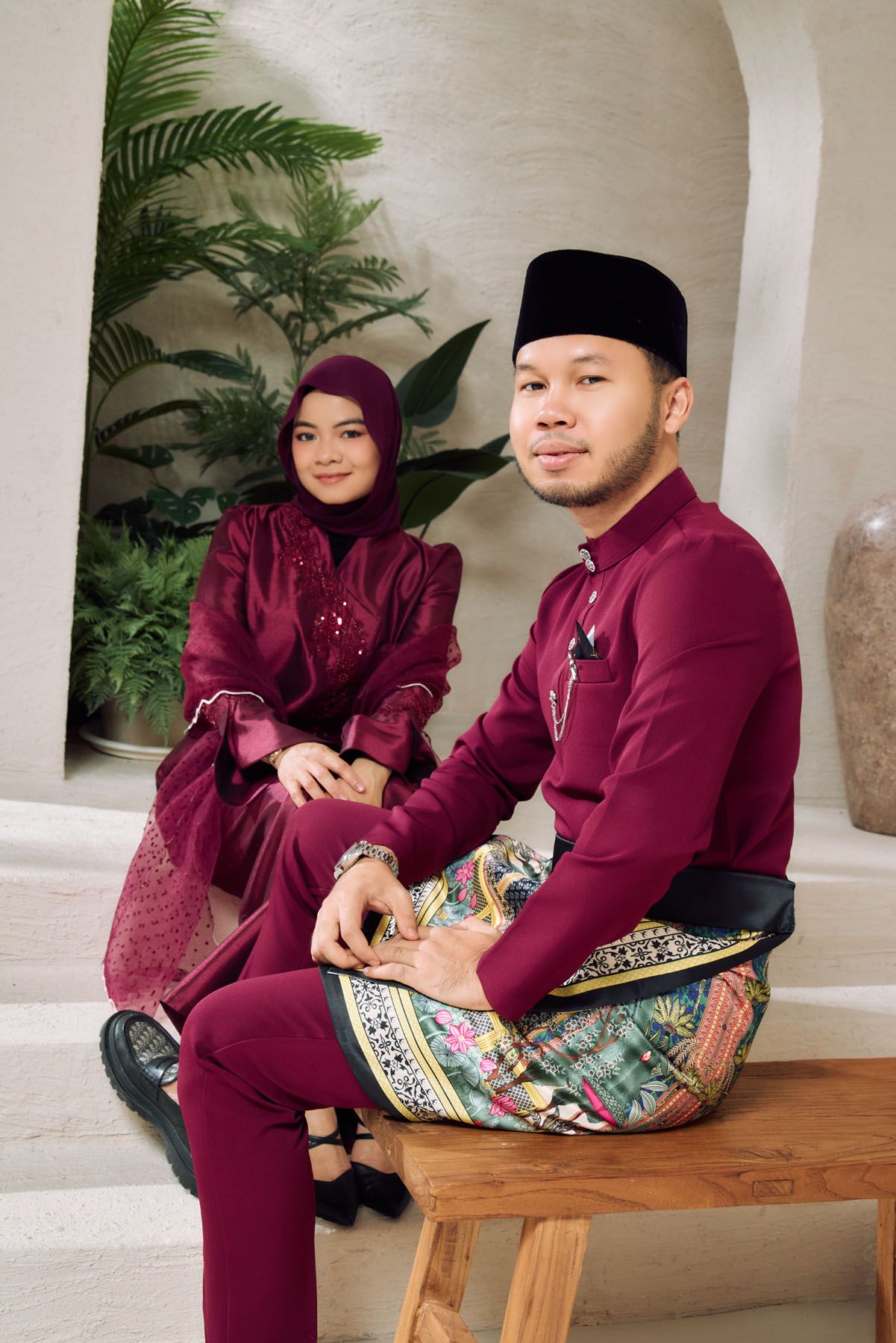 A Malaysian couple in matching burgundy traditional Baju Melayu and Baju Kurung sitting on steps in a bright interior.