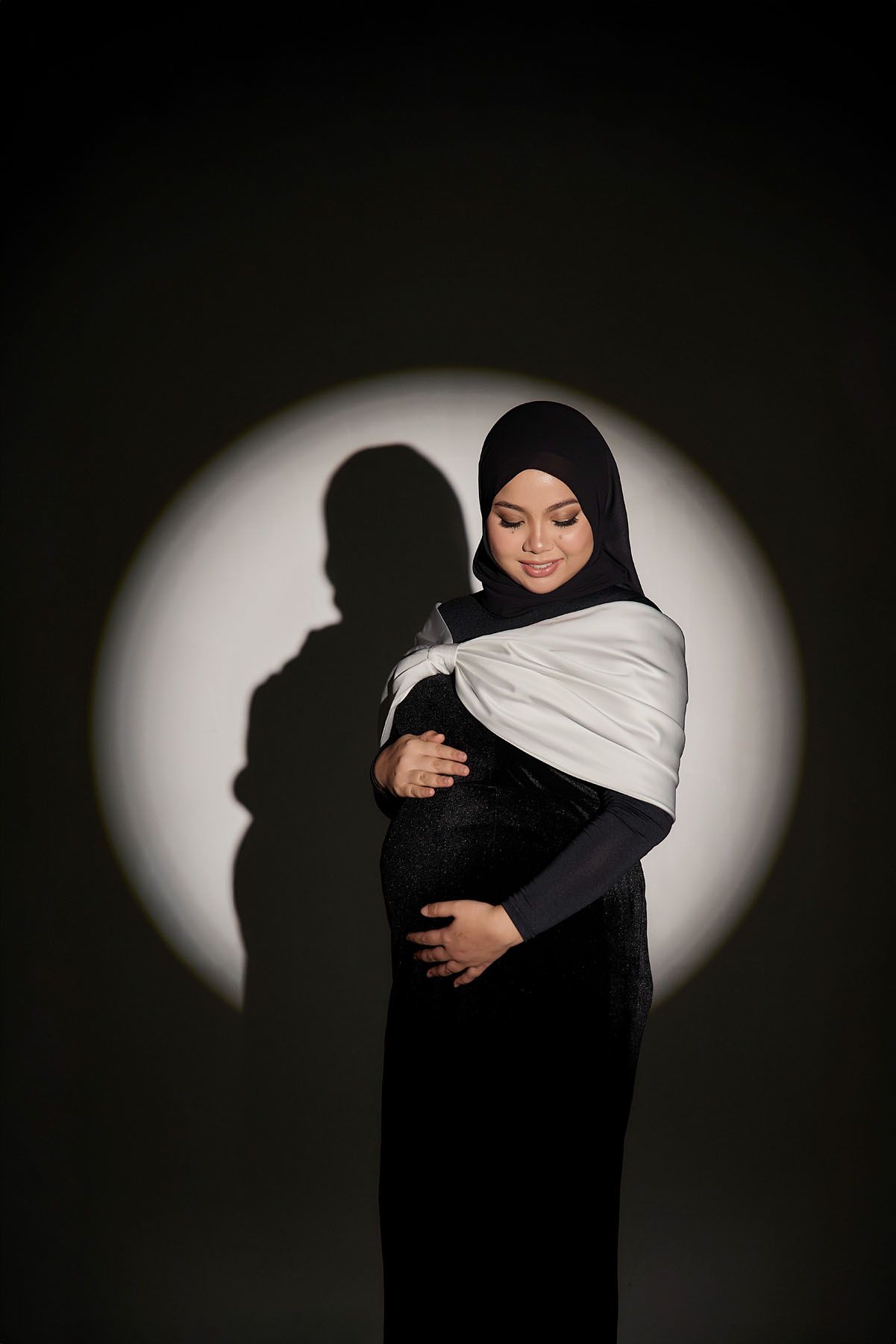 Pregnant Muslim woman in black hijab and white shawl cradling her baby bump in dramatic spotlight.