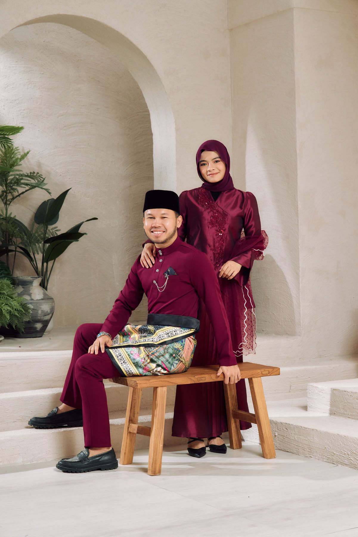 A Malaysian couple in matching maroon traditional outfits posing elegantly, man seated on bench, woman standing behind.