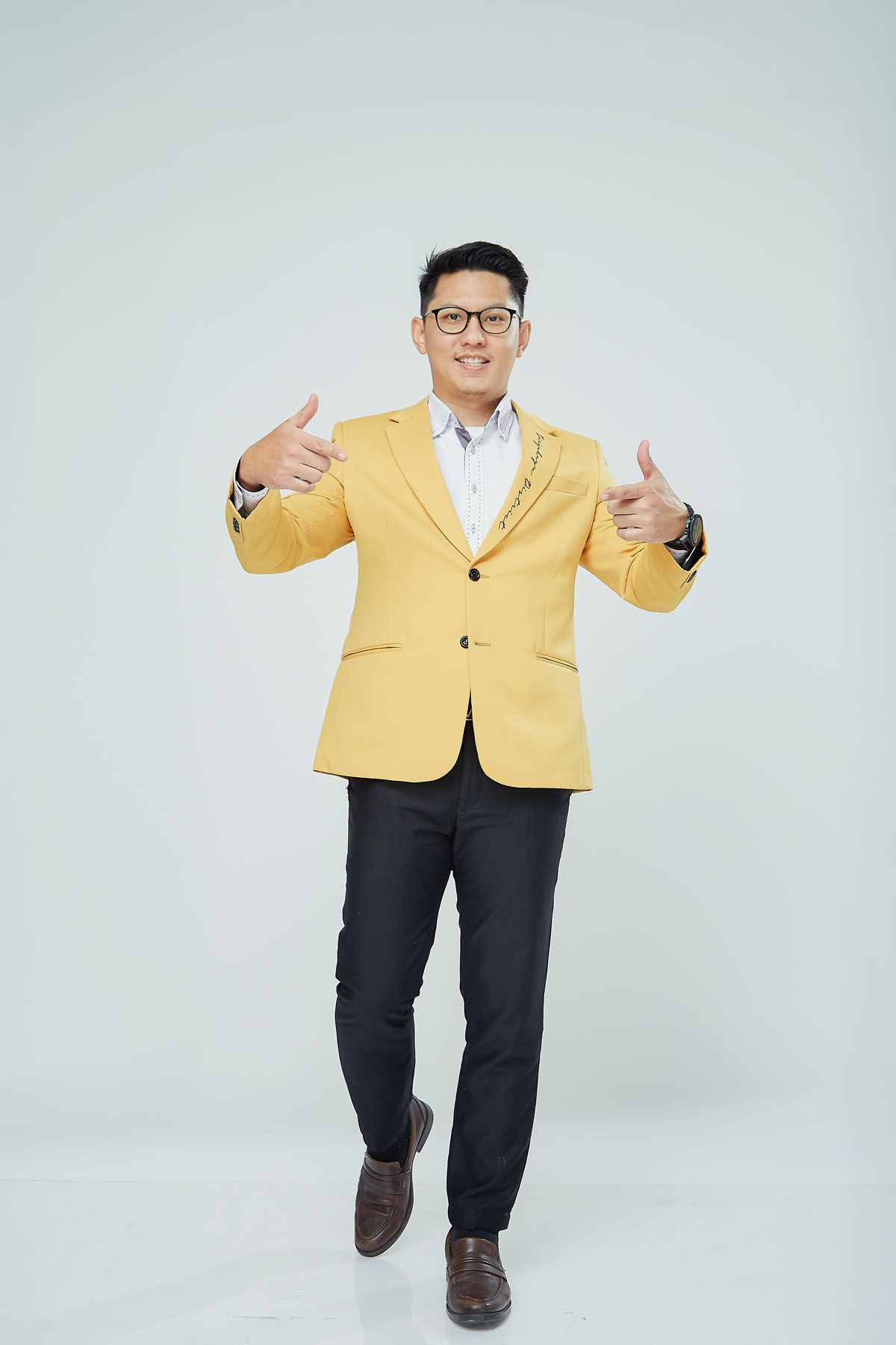 Cheerful Asian man in a yellow blazer giving two thumbs up while walking in a studio.