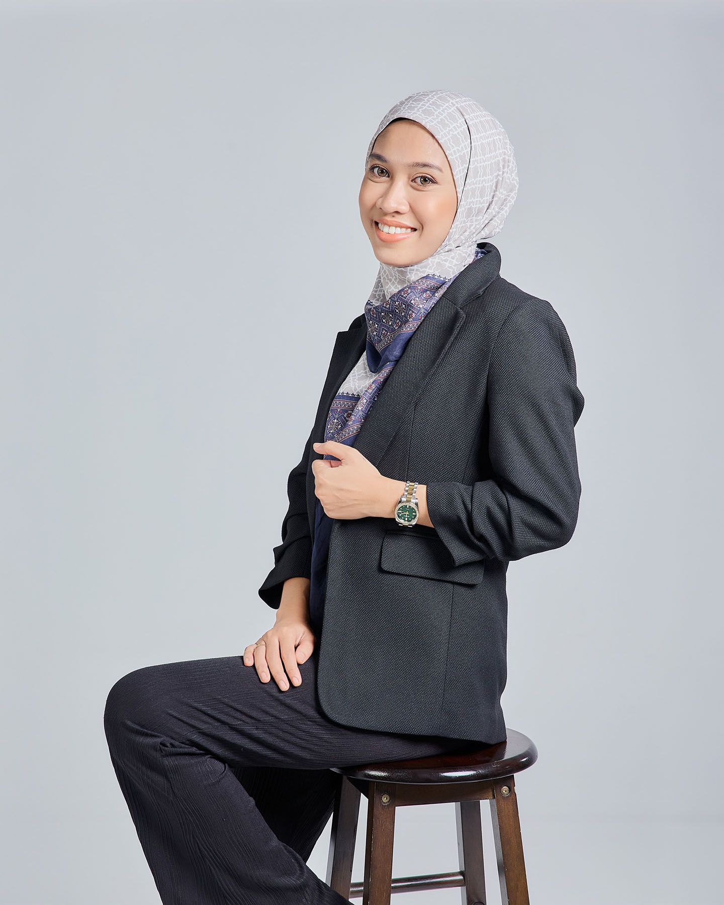 Muslim woman in dark textured blazer and patterned scarf with gray hijab smiling while seated on stool.