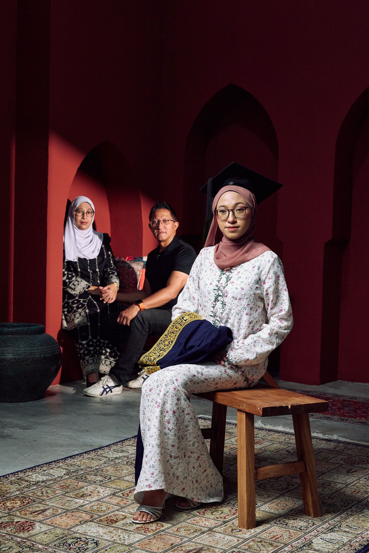 Muslim graduate in mortarboard sits on stool with parents posed behind in red archway.