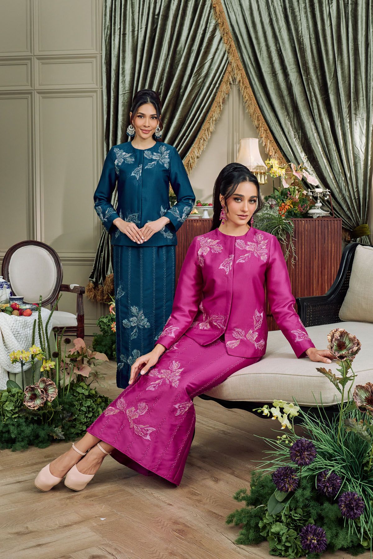 Two women in teal and magenta pink embroidered baju kurung posing in an elegant floral-decorated interior.