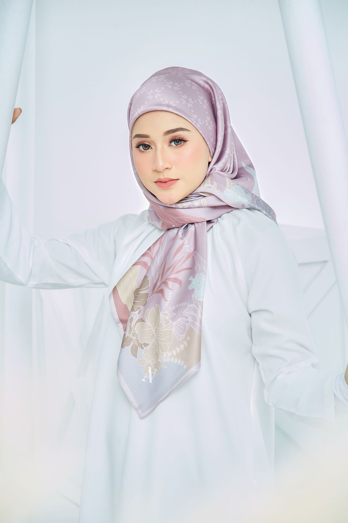 Woman modeling a lavender patterned hijab with floral accents in a soft white studio.
