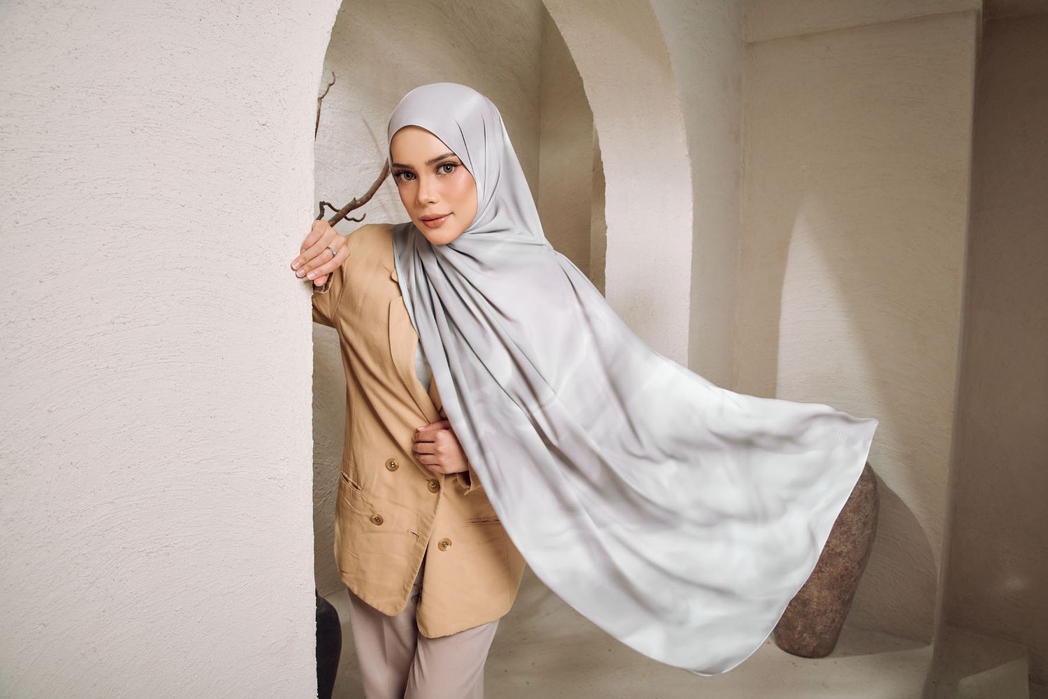 A woman in a light grey hijab and beige blazer poses against a white arched wall, holding her flowing scarf.