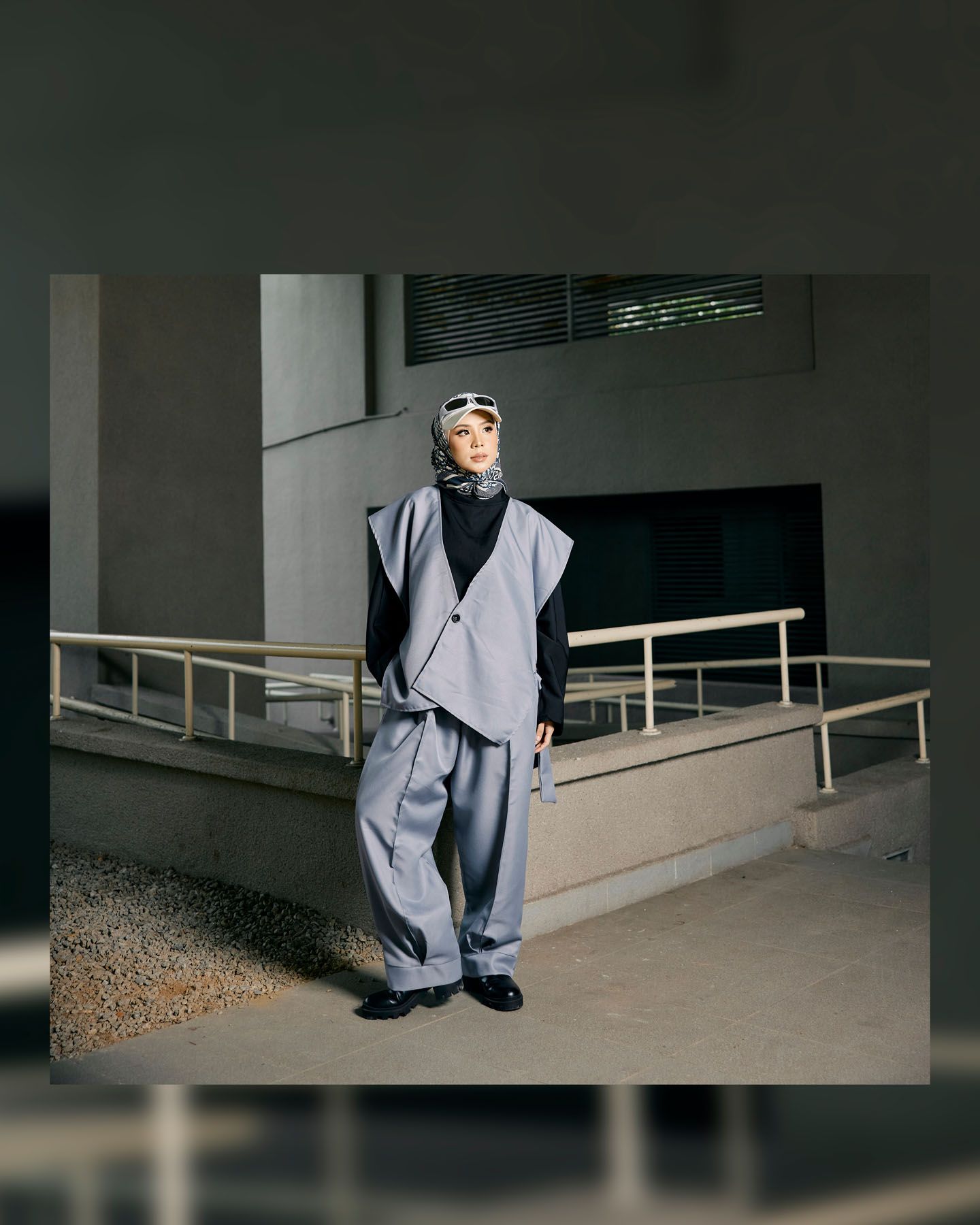 Woman in gray oversized blazer suit and hijab with sunglasses poses outdoors near modern building ramp.