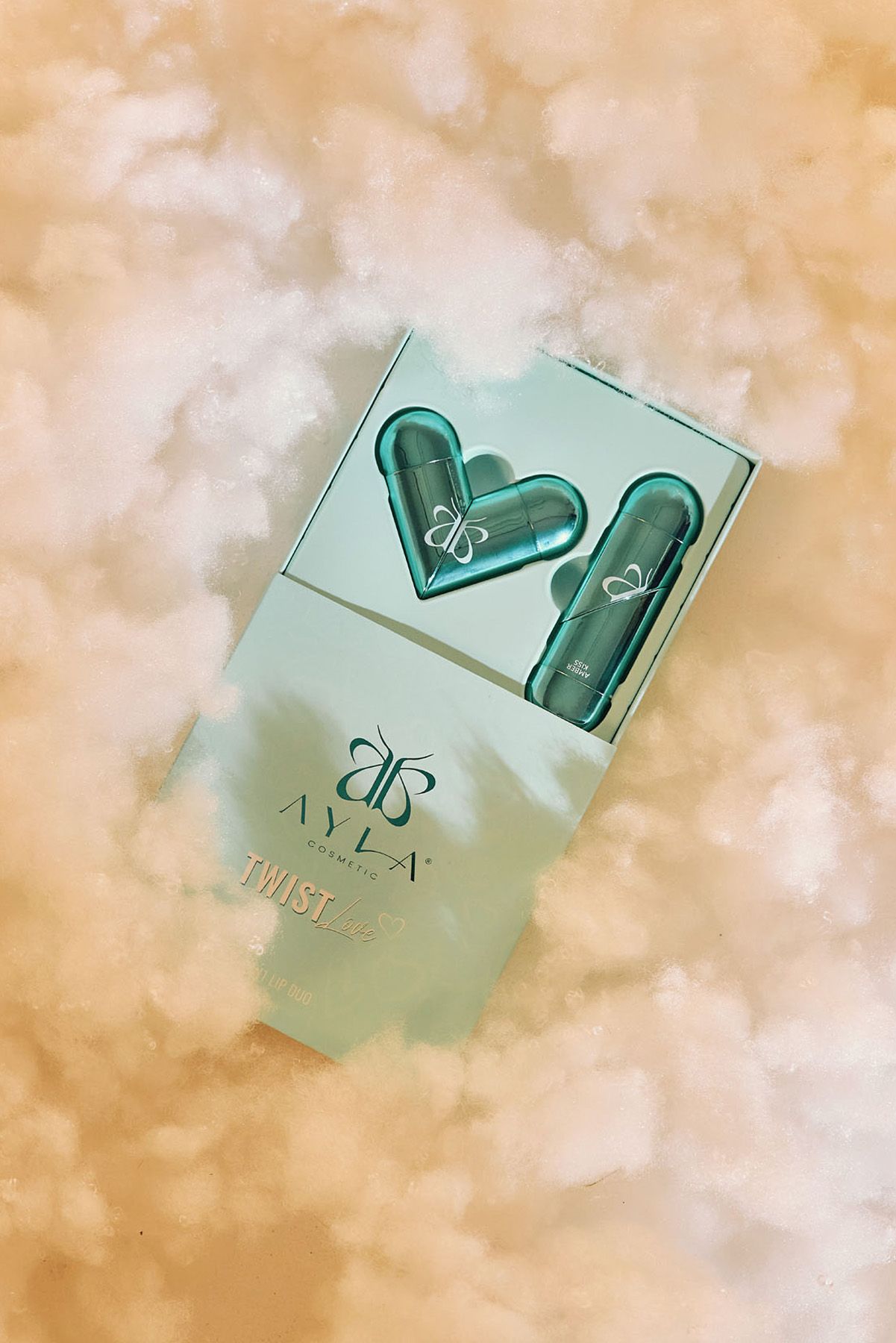 Ayla Cosmetics Twist lip gloss duo in teal packaging displayed against dreamy golden cloud background.