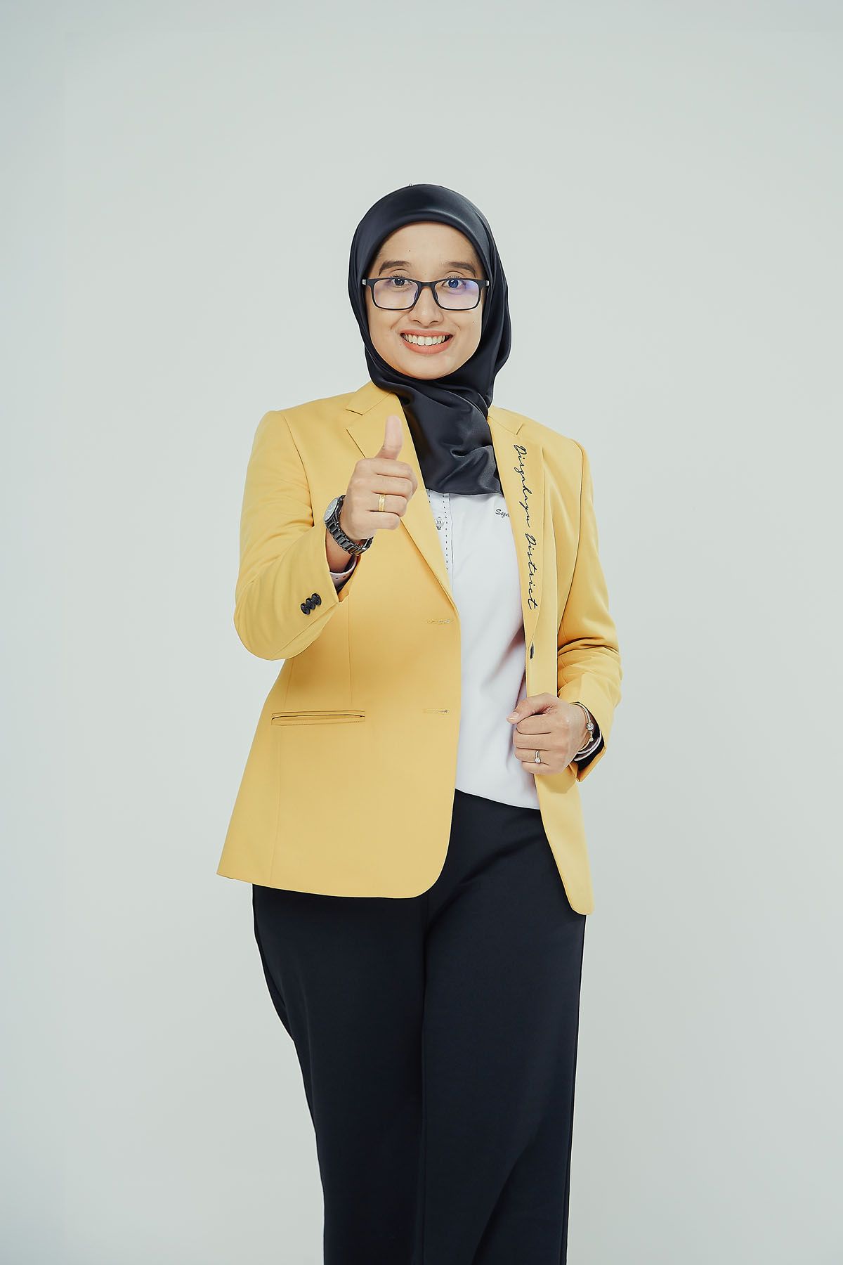 Smiling Muslim woman in a black hijab and yellow blazer pointing at camera in a studio setting.