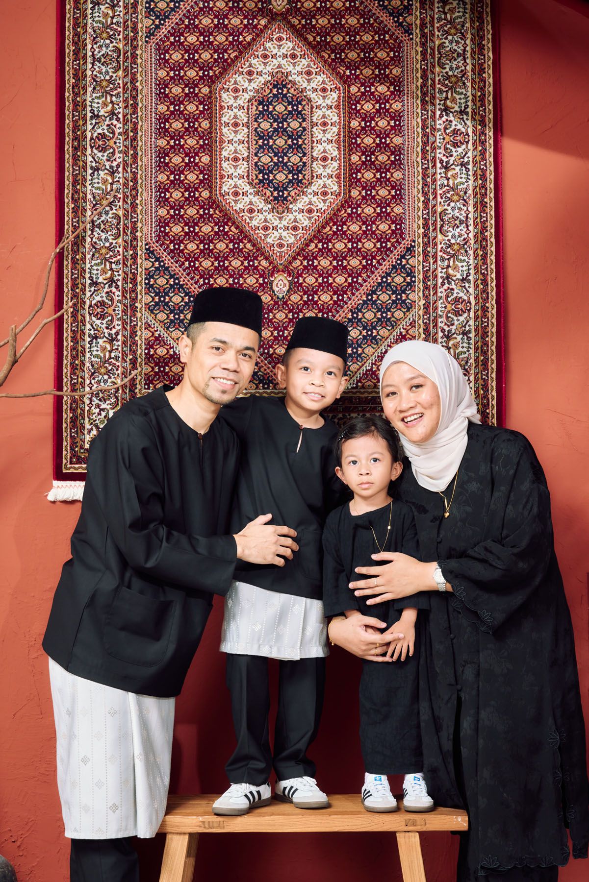 Muslim family of four in matching black baju kurung and melayu posing before red wall carpet.