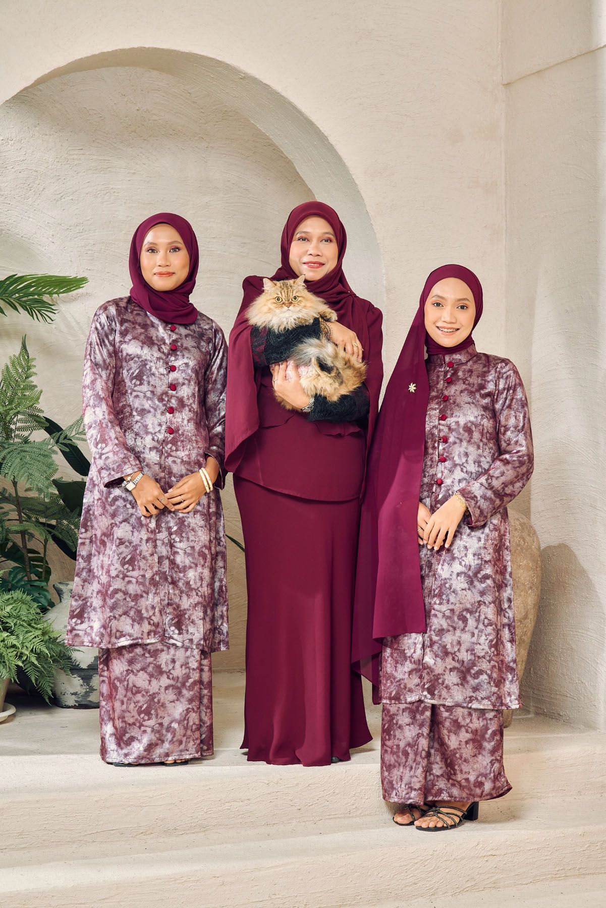 Three Muslim women in matching maroon baju kurung hijab outfits posing together, middle woman holding a fluffy cat.