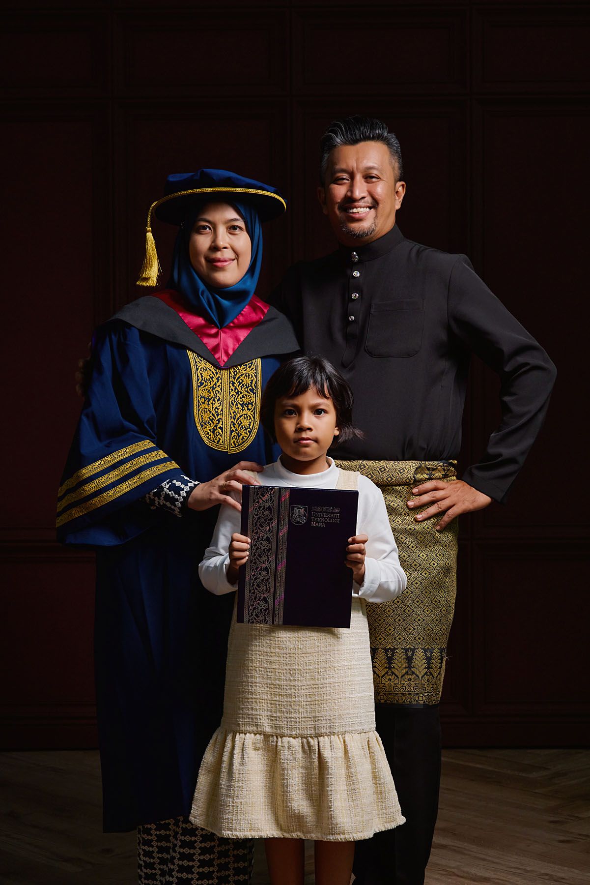 Female PhD graduate in blue doctoral robes poses with husband in black baju melayu and daughter holding thesis.