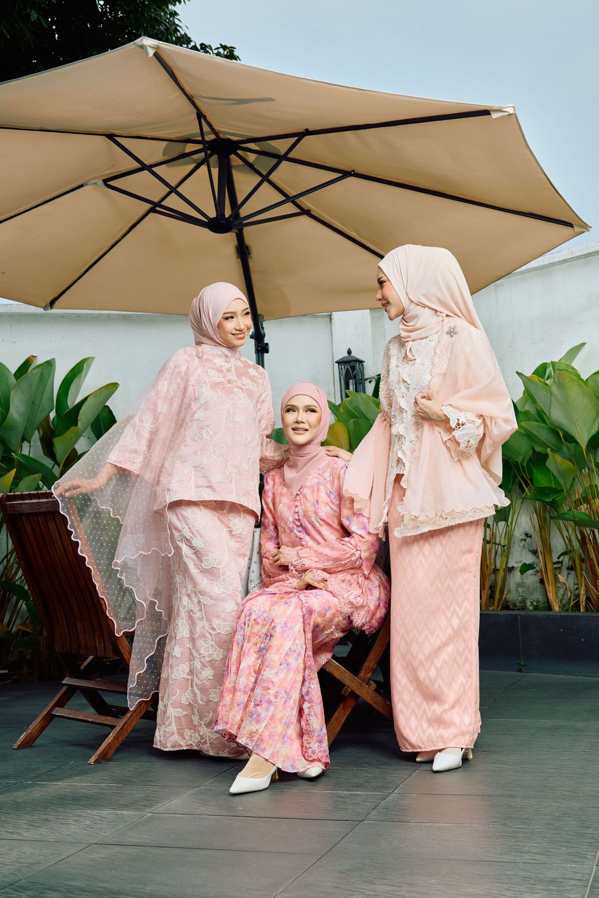 Three women in pink baju kurung and hijabs pose under a beige umbrella outdoors.