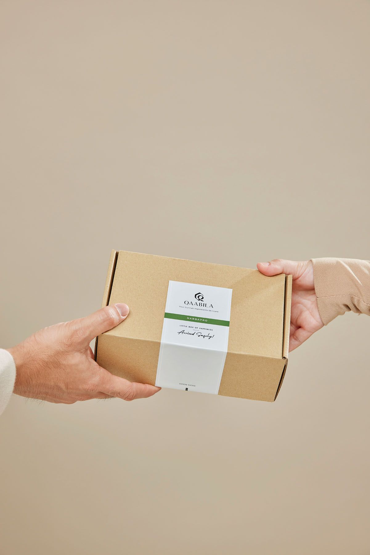 Two hands exchanging a kraft cardboard gift box with a white branded label on a neutral background.