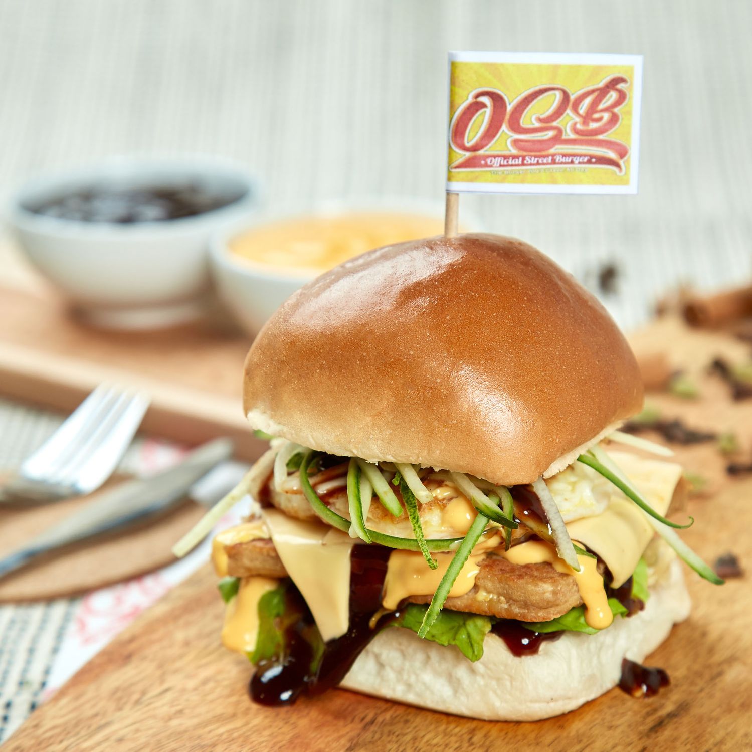 OQB branded gourmet burger with mushrooms, avocado, sprouts and BBQ sauce drizzle on wooden board.